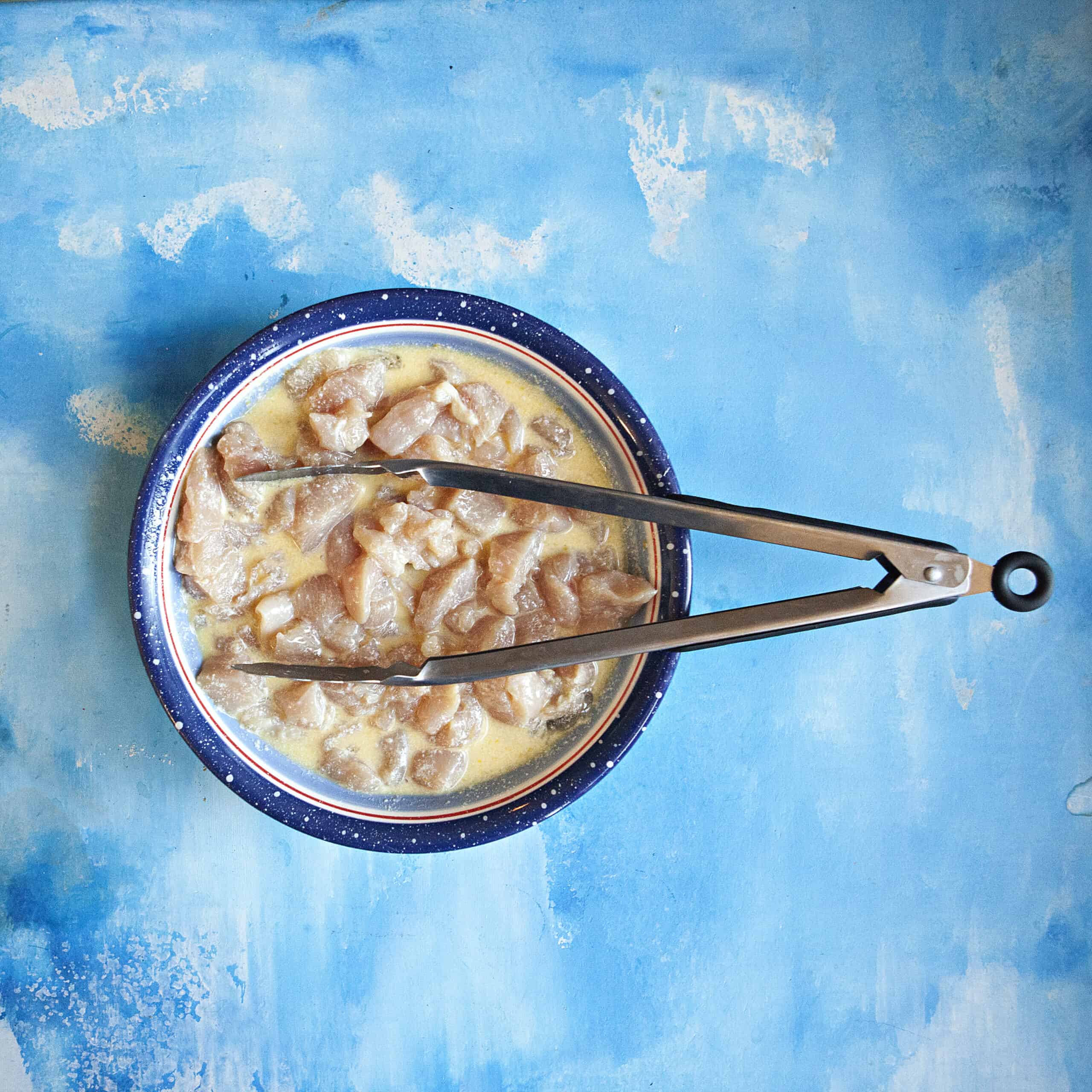 Diced raw chicken in an egg wash in a blue bowl with a pair of tongs, all on a blue backdrop.