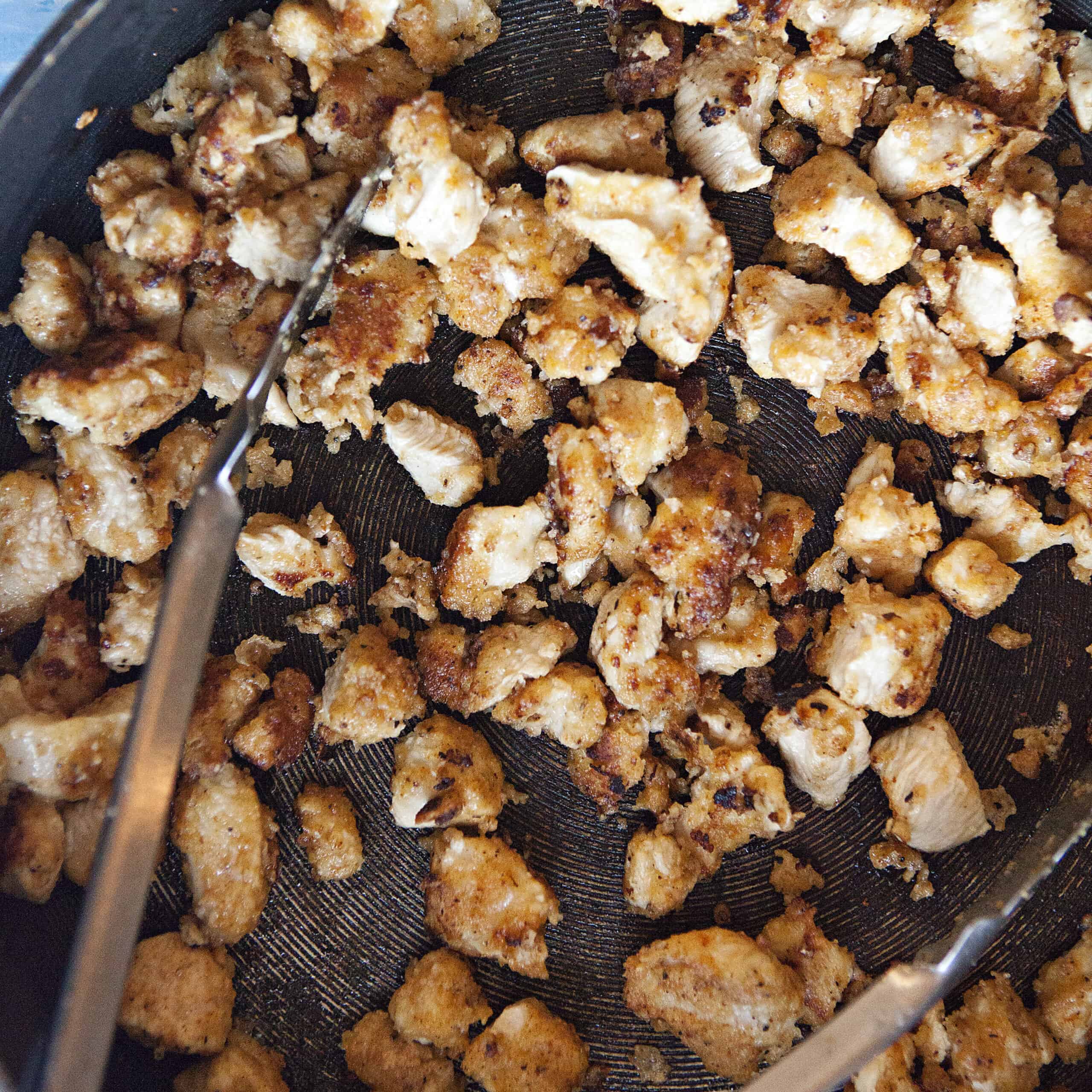 Lemon pepper chicken bites browning in a skillet with a pair of metal tongs.